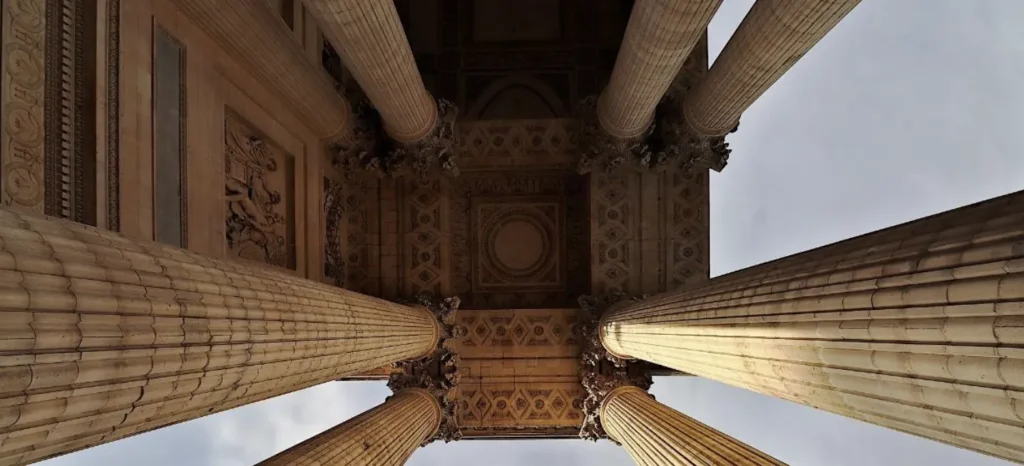View looking upward at four ornate beige stone columns supporting a detailed architectural ceiling.