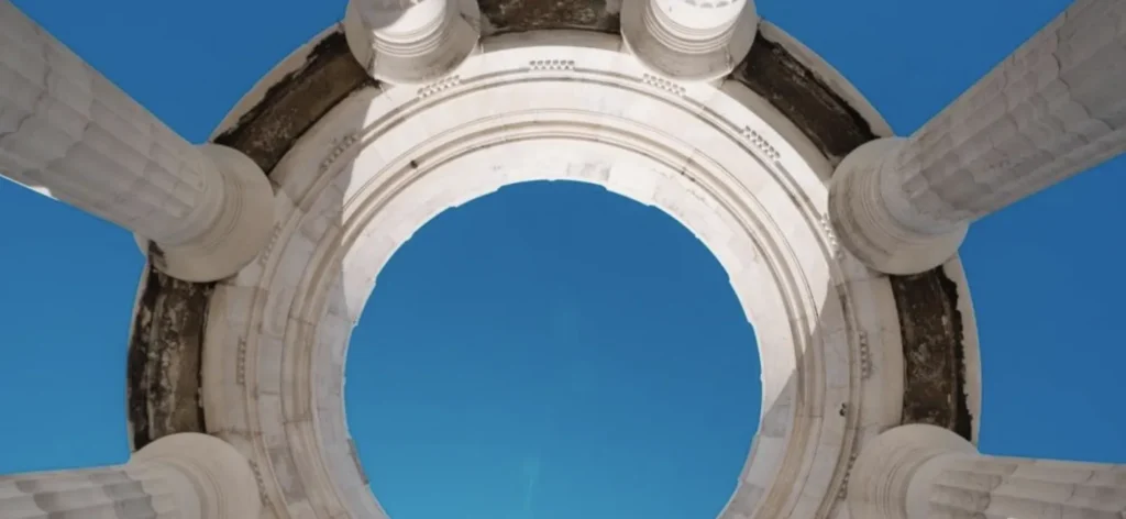 Upward view of a white circular stone arch supported by fluted columns against a clear blue sky.