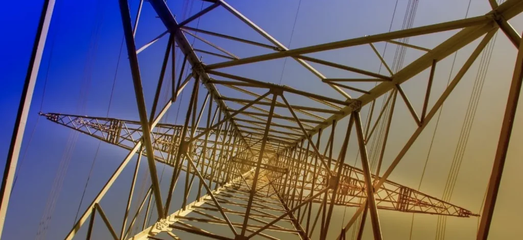 View looking upward inside a tall, yellow steel electrical transmission tower against a blue and orange gradient sky.