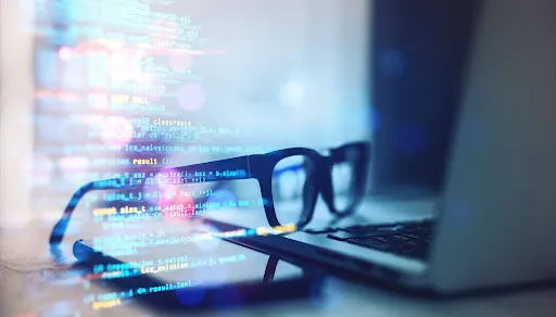Close-up of eyeglasses resting near a laptop with digital code overlay in blue hues.