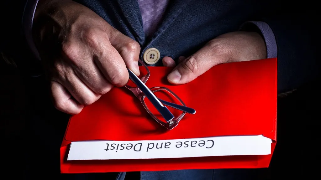 Hands holding a red folder labeled "Cease and Desist" with a pair of eyeglasses on top.