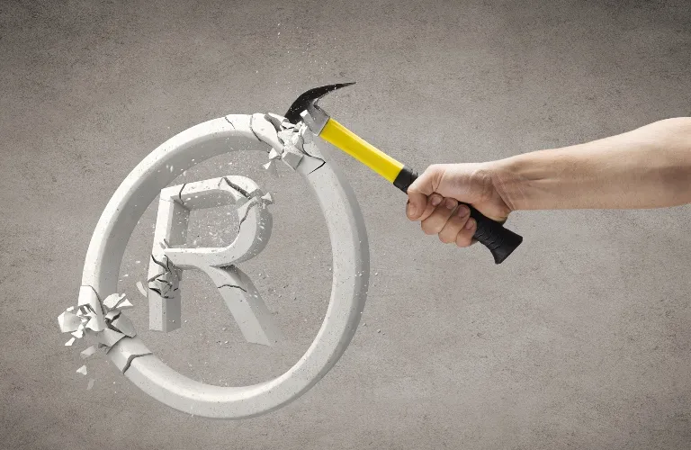 Hand holding yellow and black hammer breaking a white registered trademark symbol on gray background.