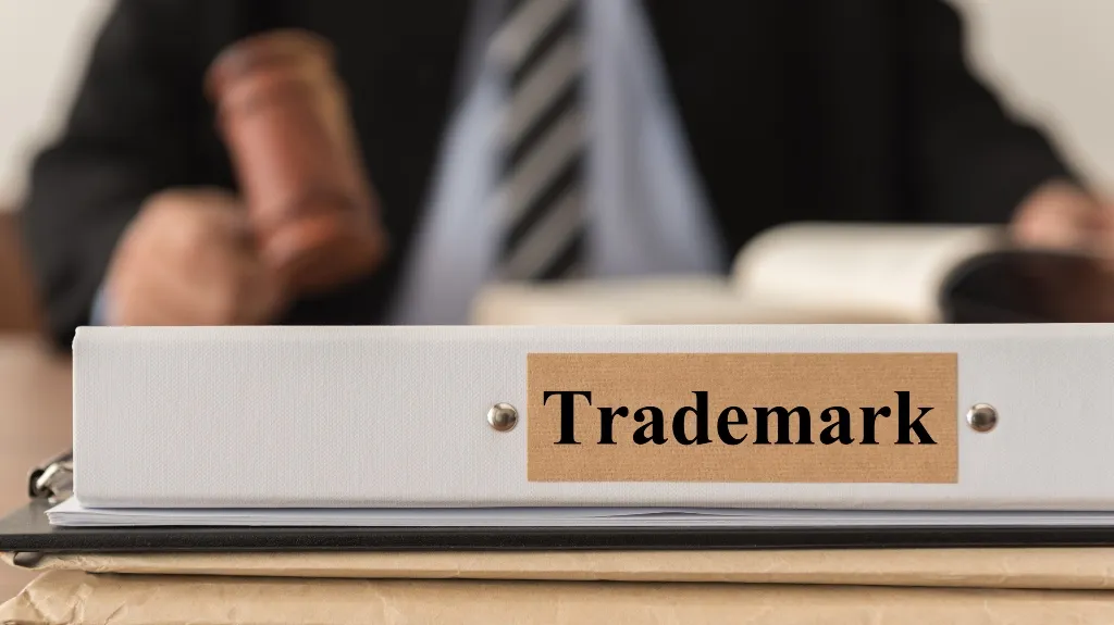 Close-up of a white binder labeled "Trademark" with a blurred person holding a gavel in the background.