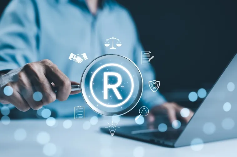 Person holding magnifying glass over registered trademark symbol near laptop, legal icons floating.