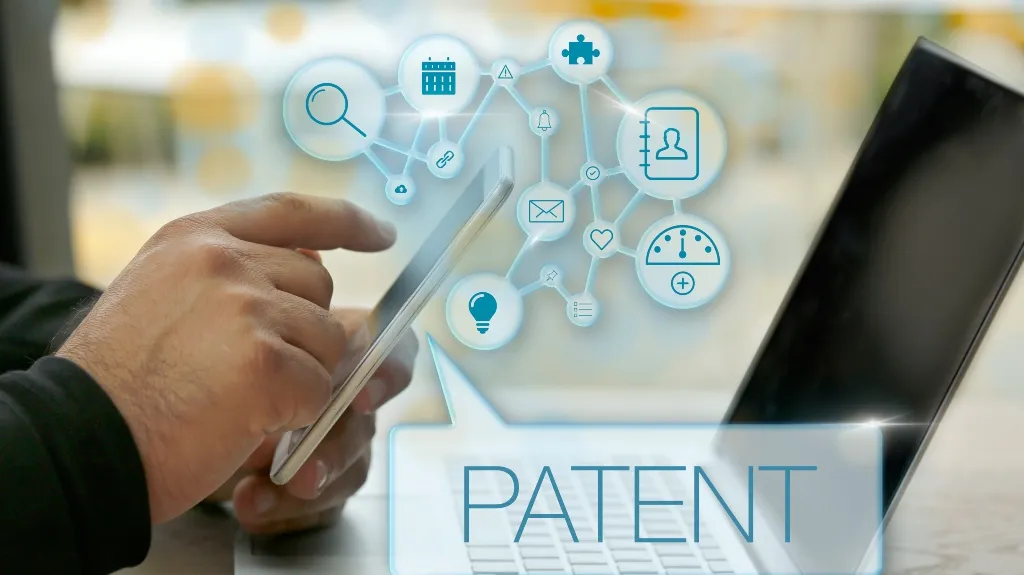 Person using a smartphone with digital icons and the word "PATENT" displayed above a laptop keyboard.